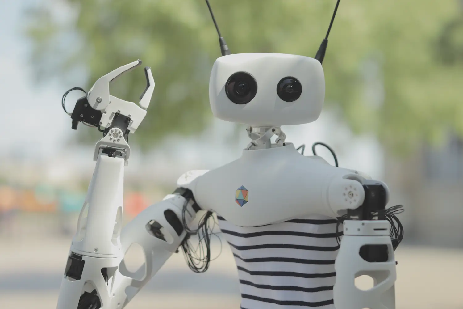 Close-up of Reachy outdoors: a white head with two camera eyes and two antennas, a navy striped shirt, one arm raised with its gripper open.
